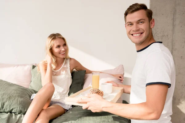 Homem sorridente trouxe café da manhã na cama para namorada em casa de manhã — Fotografia de Stock