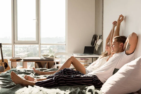 Side view of loving couple in pajamas stretching on bed at home in morning — Stock Photo