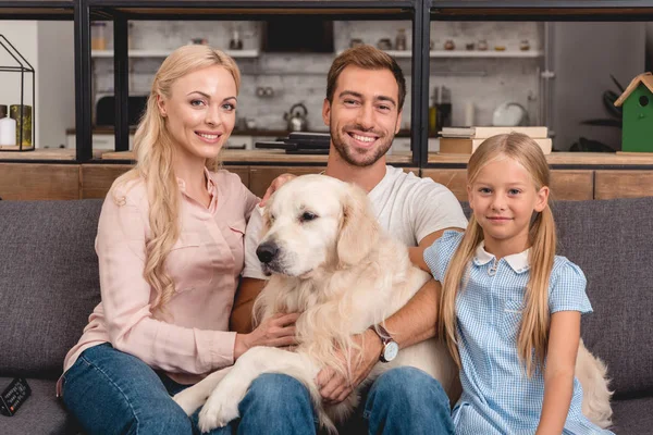 Genitori con figlia e cane seduti sul divano a casa e guardando la macchina fotografica — Foto stock