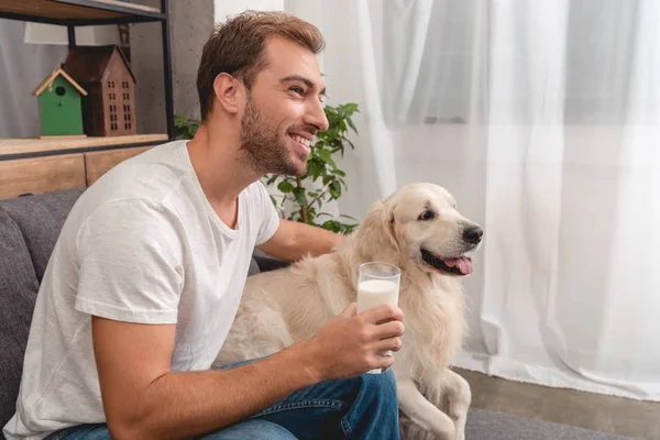 Handsome young man with glass of milk and adorable golden retriever dog sitting on couch and looking away — Stock Photo