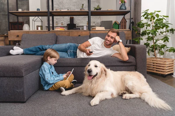 Feliz padre viendo la televisión en el sofá, mientras que su hijo usando tableta en el suelo con golden retriever - foto de stock