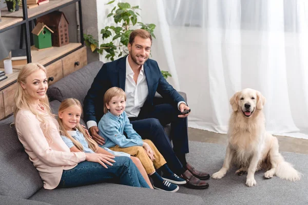 Высокий угол зрения счастливой молодой семьи, сидящей дома на диване с собакой и смотрящей в камеру — стоковое фото
