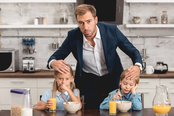 Vater überprüft Temperatur seiner kranken Kinder, indem er beim Frühstück ihre Stirn berührt und in die Kamera schaut — Stockfoto