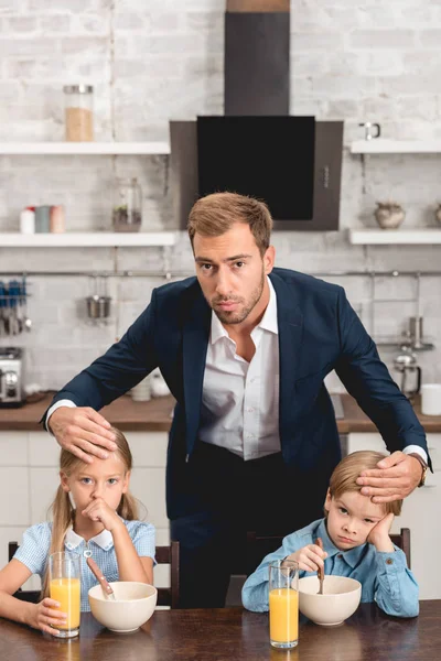 Young father checking temperature of his sick kids by touching their foreheads during breakfast and looking at camera — Stock Photo
