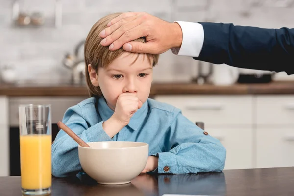 Tiro recortado de padre comprobar la temperatura del hijo con la mano durante el desayuno - foto de stock