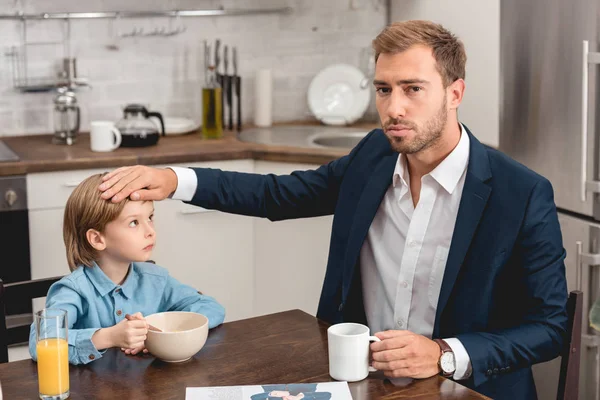 Грустный отец проверяет температуру сына с руки во время завтрака — стоковое фото