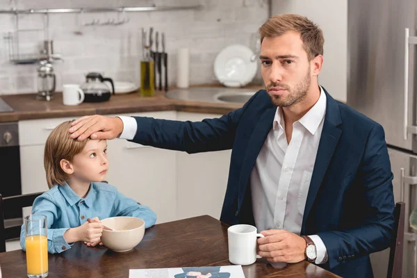 Padre triste comprobar la temperatura del hijo con la mano durante el desayuno - foto de stock