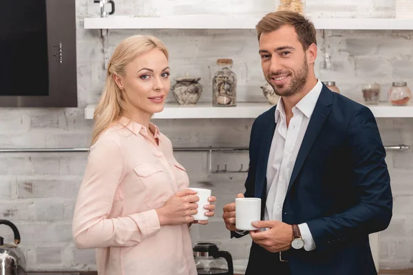 Feliz pareja sosteniendo tazas de café y mirando a la cámara en la cocina - foto de stock