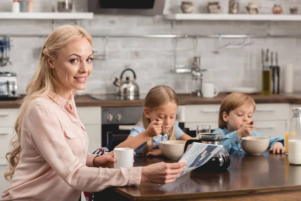 Усміхнена мати має чашку кави, сидячи на кухні з дітьми під час сніданку і дивлячись на камеру — стокове фото