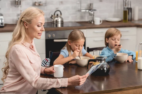 Attractive mother having cup of coffee while sitting at kitchen with kids during breakfast — Stock Photo
