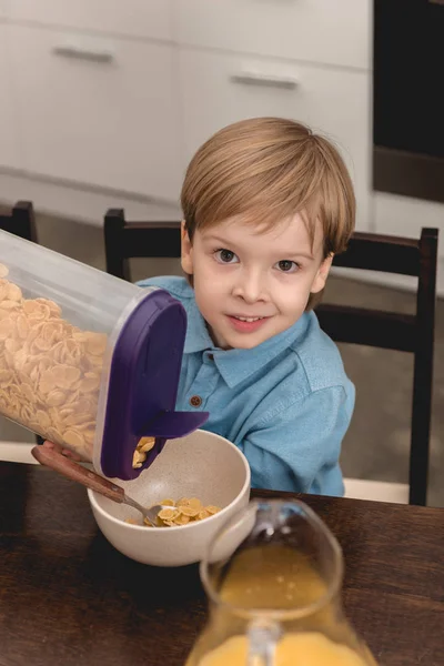 Entzückendes kleines Kind schüttet Müsli in Schüssel zum Frühstück und schaut in die Kamera — Stockfoto