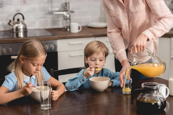 Plan recadré de mère versant du jus d'orange pour les enfants pendant le petit déjeuner — Photo de stock