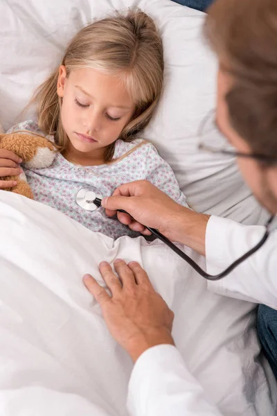 Pediatra confiado litening niños respiración con estetoscopio mientras ella acostada en la cama - foto de stock