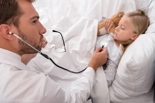 High angle view of confident pediatrician litening childs breath with stethoscope while she lying in bed — Stock Photo
