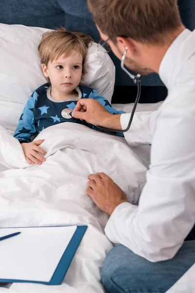 High angle view of pediatrician litening childs breath with stethoscope while he lying in bed — Stock Photo