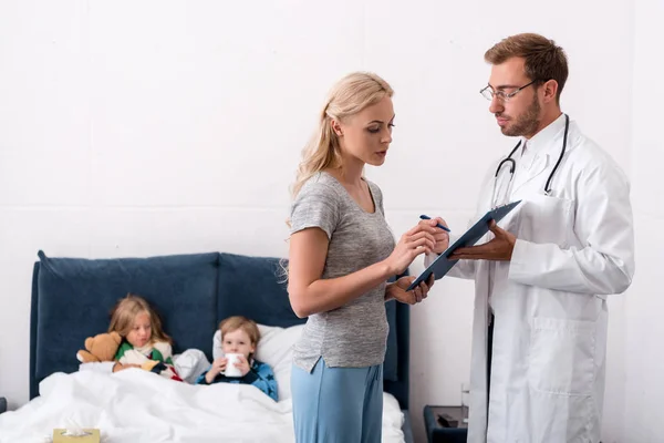 Pediatrician giving clipboard to mother of diseased kids lying in bed on background to sign documents — Stock Photo