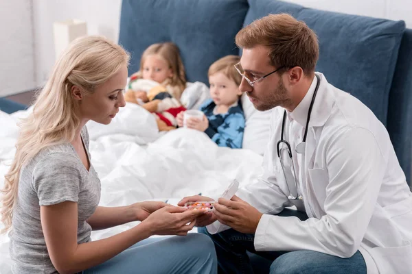 Pediatrician giving pills to mother of diseased kids lying in bed on background — Stock Photo