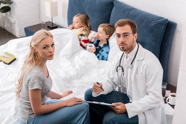 Vista de ángulo alto del pediatra y la madre mirando a la cámara mientras los niños enfermos que yacen en la cama sobre el fondo - foto de stock