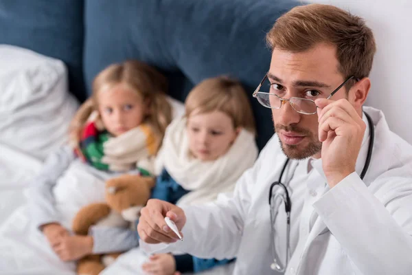 Vista de ángulo alto del hermoso pediatra con termómetro mirando a la cámara mientras está sentado cerca de los niños acostados en la cama - foto de stock