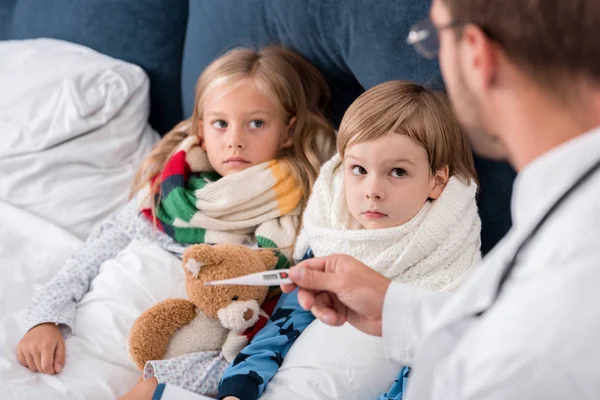 Junge Kinderärztin mit Klemmbrett untersucht Temperatur von im Bett liegenden Kindern — Stockfoto