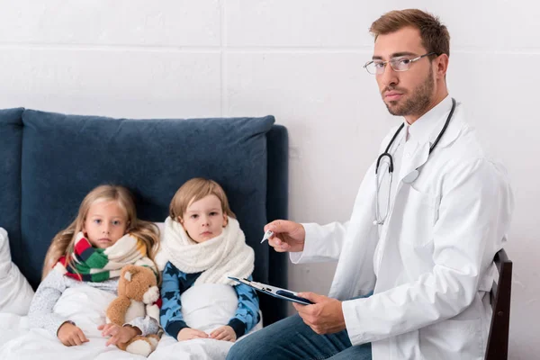Kinderarzt schreibt in Klemmbrett und blickt in die Kamera, während er neben Kindern sitzt, die im Bett liegen — Stockfoto