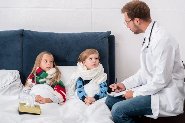 Schöner junger Kinderarzt mit Klemmbrett, der auf im Bett liegende Kinder blickt — Stockfoto