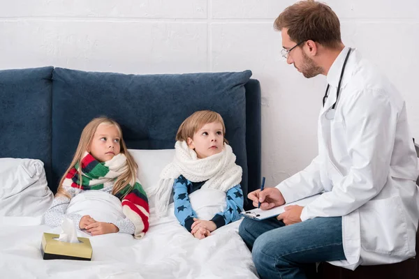 Beau jeune pédiatre avec presse-papiers regardant les enfants couchés dans le lit — Photo de stock