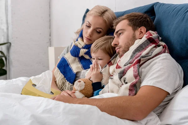 Krankes kleines Kind pustet Nase, während es mit Eltern im Bett liegt — Stockfoto