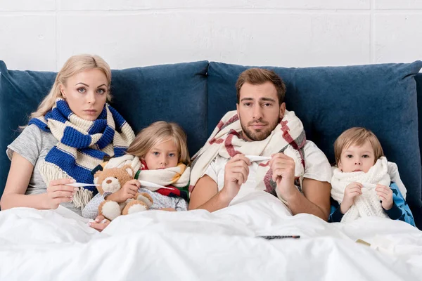 Kranke junge Familie in Schals hält elektrische Thermometer in der Hand und blickt in die Kamera, während sie im Bett liegt — Stockfoto