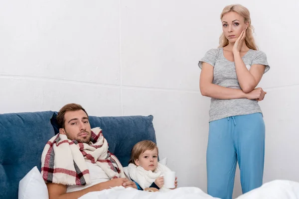 Confused woman standing near sick husband and son and looking at camera while they lying in bed — Stock Photo