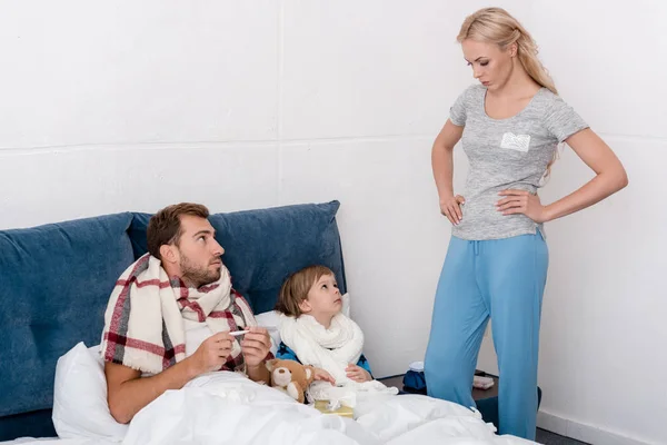 Mujer de pie cerca de marido enfermo e hijo mientras yacen en la cama - foto de stock