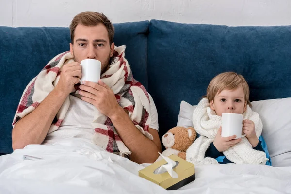 Kranker Vater und Sohn in Schals, die Tee trinken, während sie im Bett sitzen und in die Kamera schauen — Stockfoto