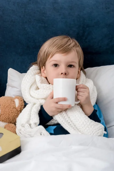 Sick little child in scarf with with cup of tea lying in bed and looking at camera — Stock Photo