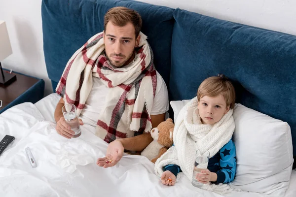 Vista ad alto angolo di padre e figlio malato con bicchieri d'acqua e pillole seduti a letto e guardando la fotocamera — Foto stock