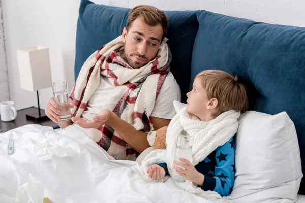 Padre e hijo enfermos con vasos de agua y pastillas sentados en la cama y mirándose el uno al otro - foto de stock