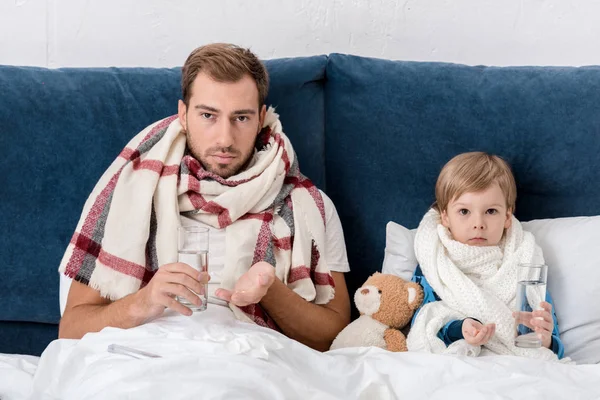 Sick father and son with glasses of water and pills sitting in bed and looking at camera — Stock Photo