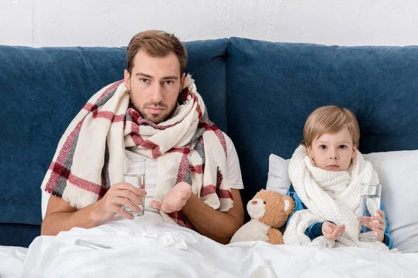 Padre e hijo enfermos con vasos de agua y pastillas sentados en la cama y mirando a la cámara - foto de stock