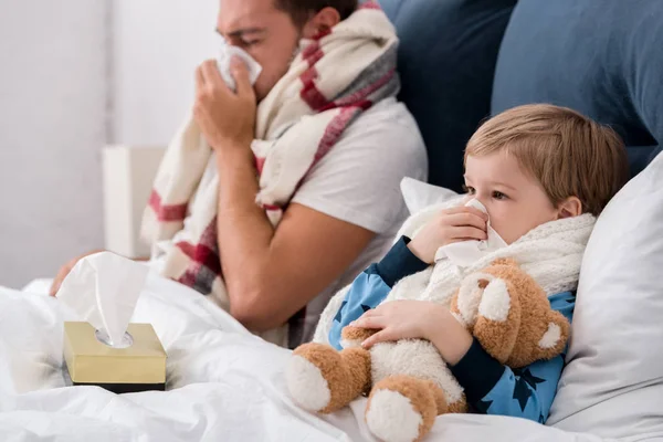 Malade père et fils soufflant nez avec des serviettes en papier tout en étant couché dans le lit — Photo de stock