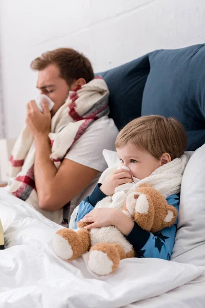Jeune père et fils soufflant nez avec des serviettes en papier tout en étant couché dans le lit — Photo de stock