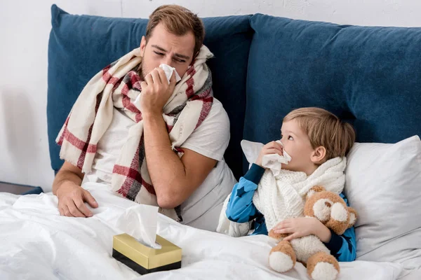 Father and son blowing noses with paper napkins while lying in bed and looking at each other — Stock Photo