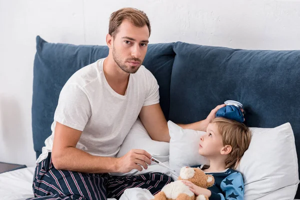 Padre comprobando la temperatura del hijo con termómetro eléctrico y sosteniendo la bolsa de hielo en su cabeza en la cama y mirando a la cámara - foto de stock