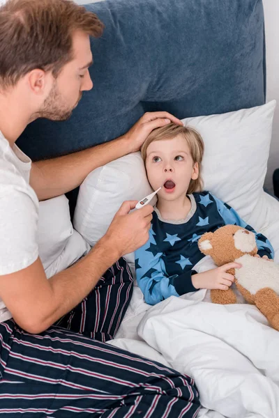 Visão de alto ângulo do pai verificando a temperatura oral do filho enquanto ele deitado na cama — Fotografia de Stock