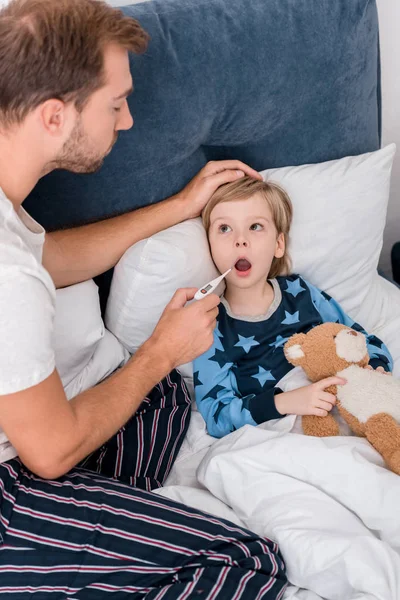 Vue grand angle du père vérifiant la température orale du fils alors qu'il était couché au lit — Photo de stock