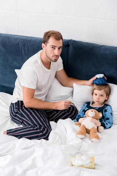 High angle view of father checking temperature of son with electric thermometer and holding ice pack on his head in bed and looking at camera — Stock Photo