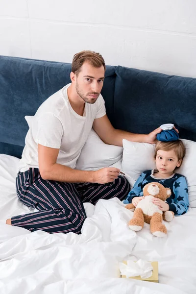 Vista de ángulo alto del padre comprobando la temperatura del hijo con termómetro eléctrico y sosteniendo la bolsa de hielo en su cabeza en la cama y mirando a la cámara - foto de stock