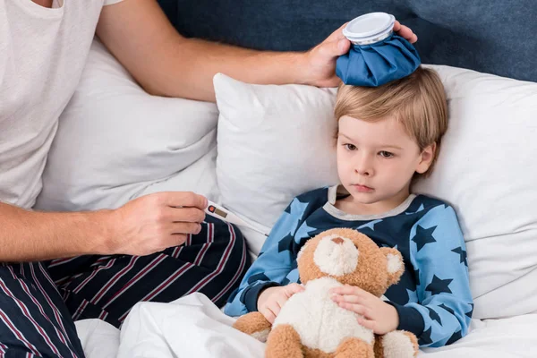 Recortado disparo de padre comprobando temperatura de hijo con termómetro eléctrico y sosteniendo bolsa de hielo en su cabeza en la cama - foto de stock