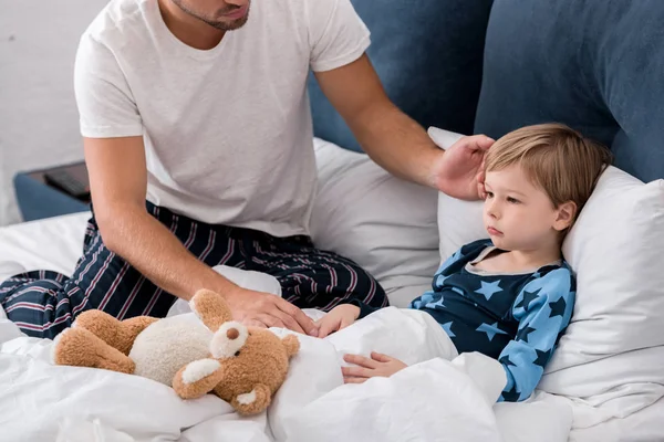 Recortado disparo de joven padre comprobar hijos temperatura con la mano mientras que él acostado en la cama - foto de stock