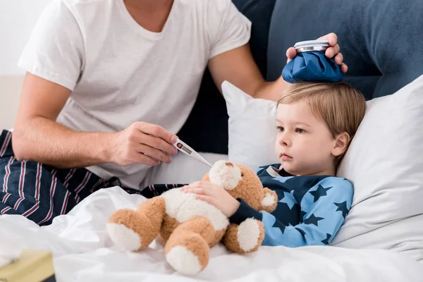 Cropped shot of father checking sons temperature with electric thermometer and holding ice pack on his head in bed — Stock Photo