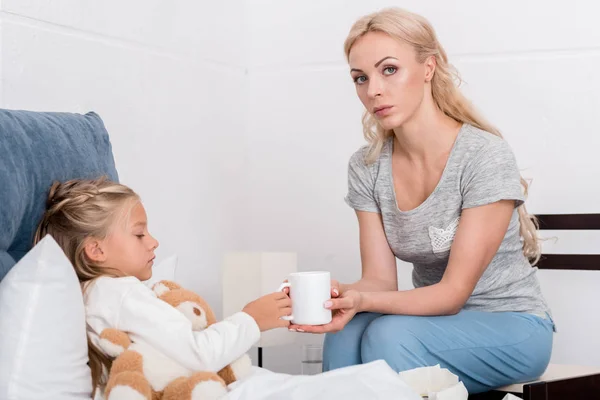 Joven madre dando taza de té caliente a su hija enferma en la cama - foto de stock