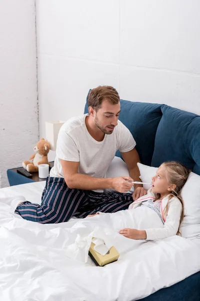 Молодий батько перевіряє дочок на пероральну температуру, поки вона лежить у ліжку — стокове фото